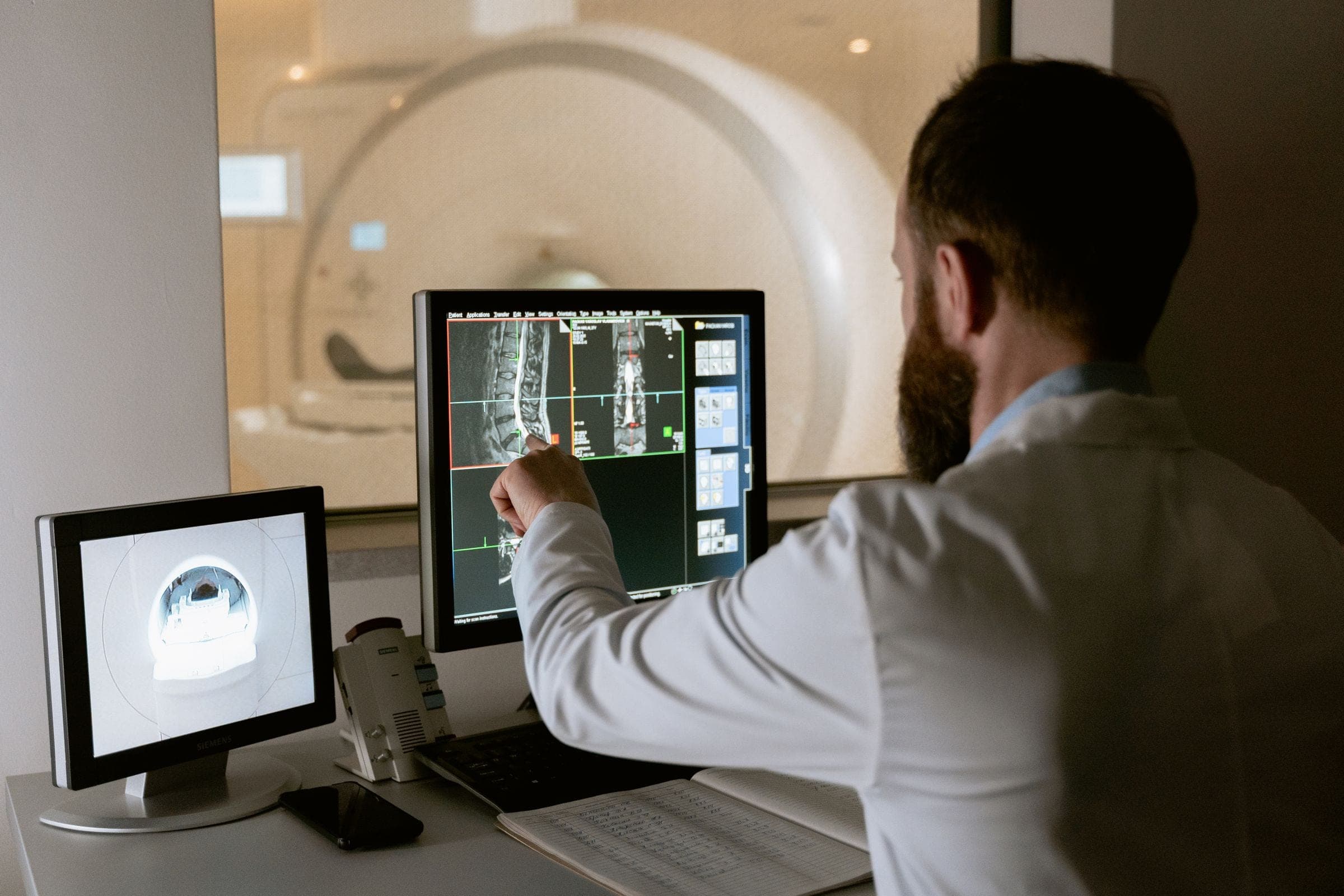 A clinician reviewing a study on a reporting workstation, the scanner visible through the window beyond.