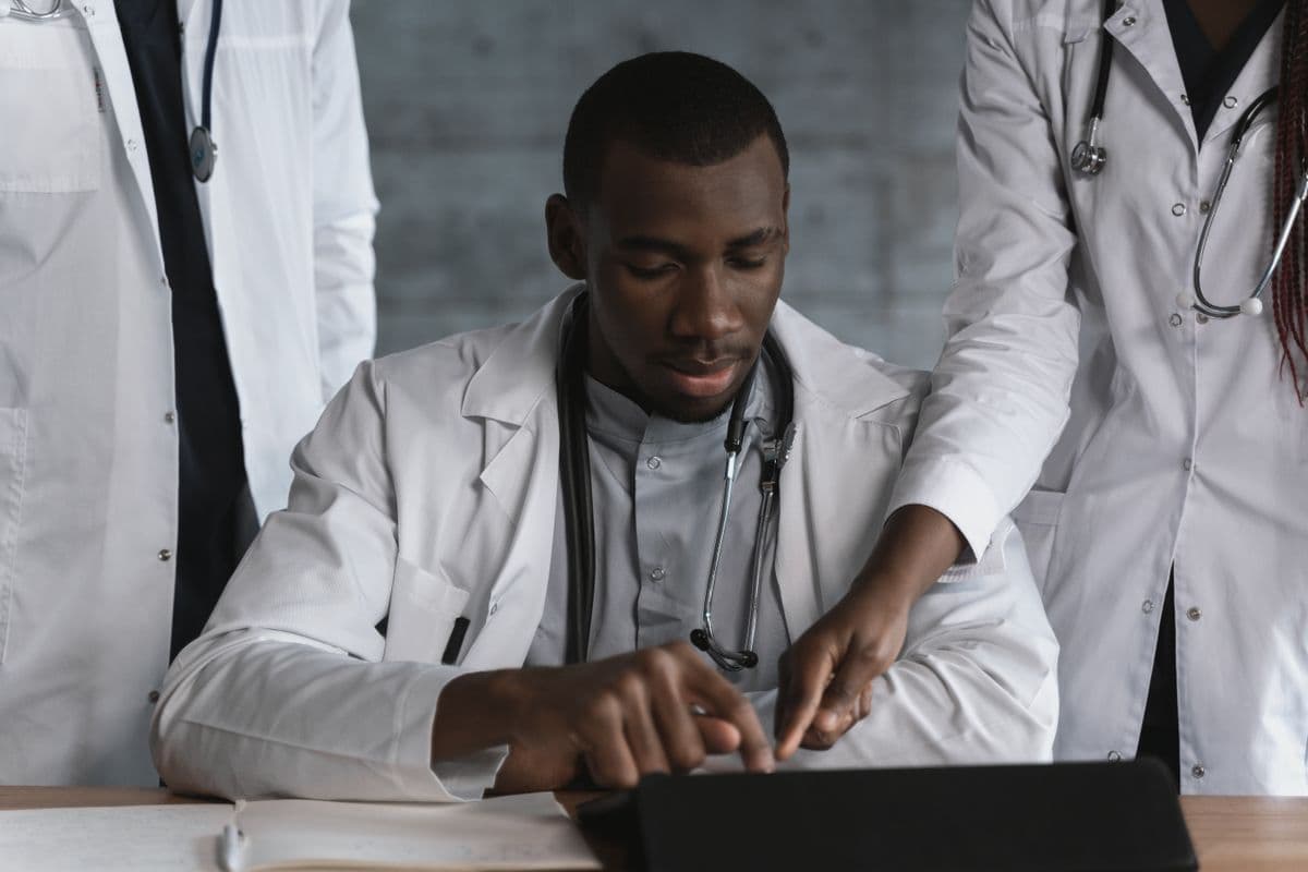 A clinician in a white coat reviewing work at a laptop with colleagues in a clinical setting.